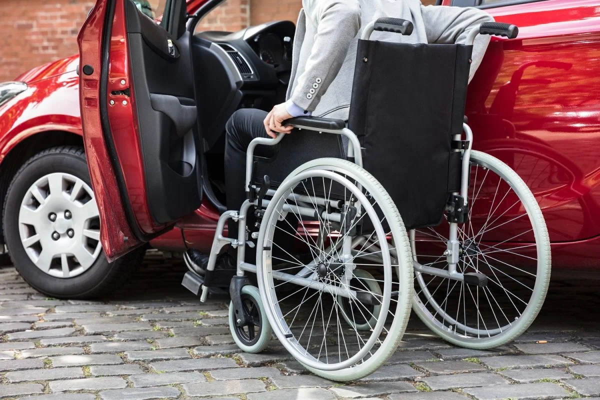 Pessoa em cadeira de rodas ao lado de carro adaptado, relacionada ao tema PCD.